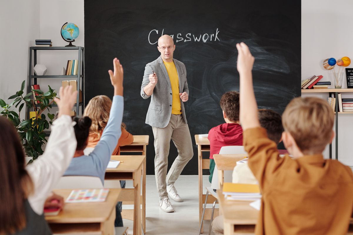 A teacher at the front of a classroom with students raising their hands